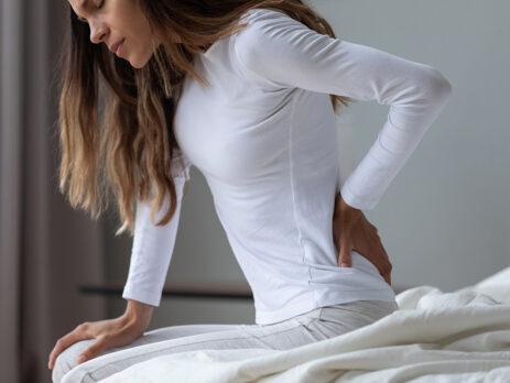 woman holding his back because of pain while sitting on a bedside