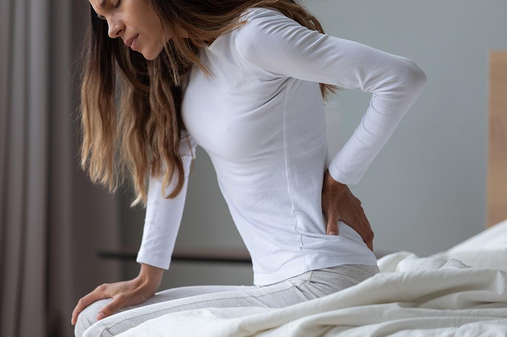 woman holding his back because of pain while sitting on a bedside